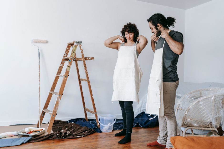 A couple prepares for home renovation, wearing aprons in an indoor setting.