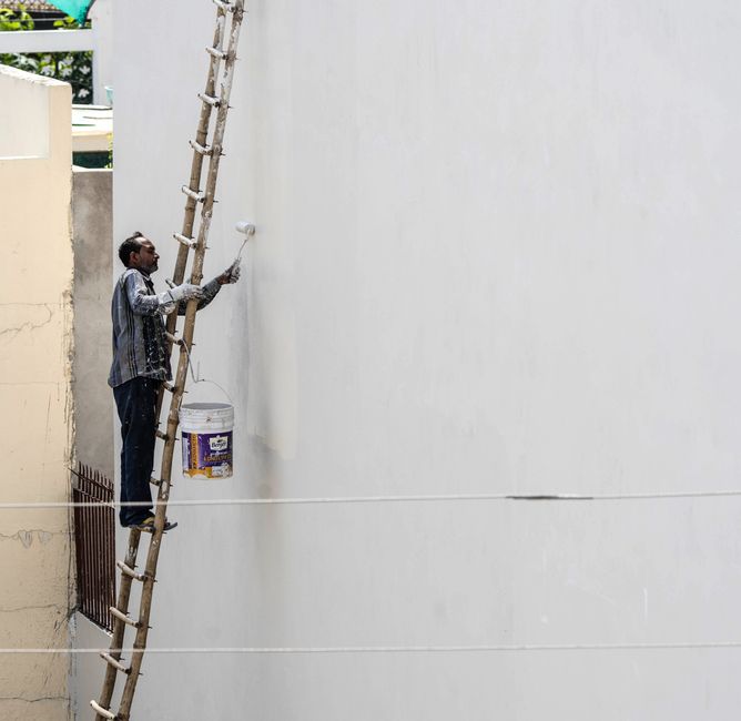 A man on a ladder painting a building's exterior wall with a roller brush outdoors.