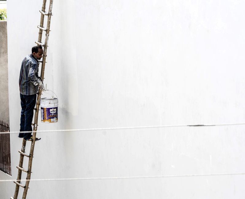A worker on a ladder paints a large white wall outdoors, showcasing construction work.